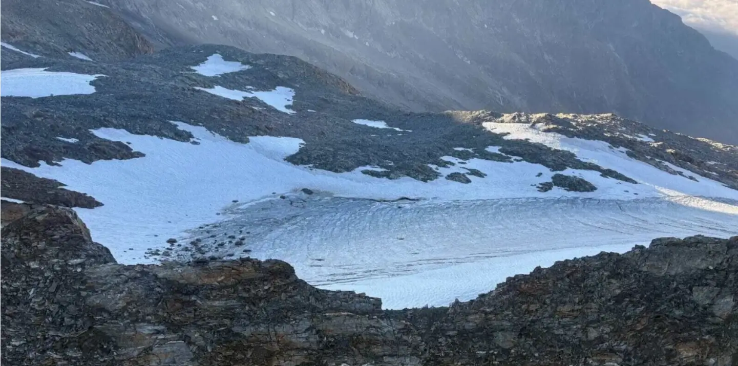 A scenic view of a snowy mountain landscape with patches of snow and rocky terrain, featuring a glacier or snowfield nestled between rugged peaks under a cloudy sky.