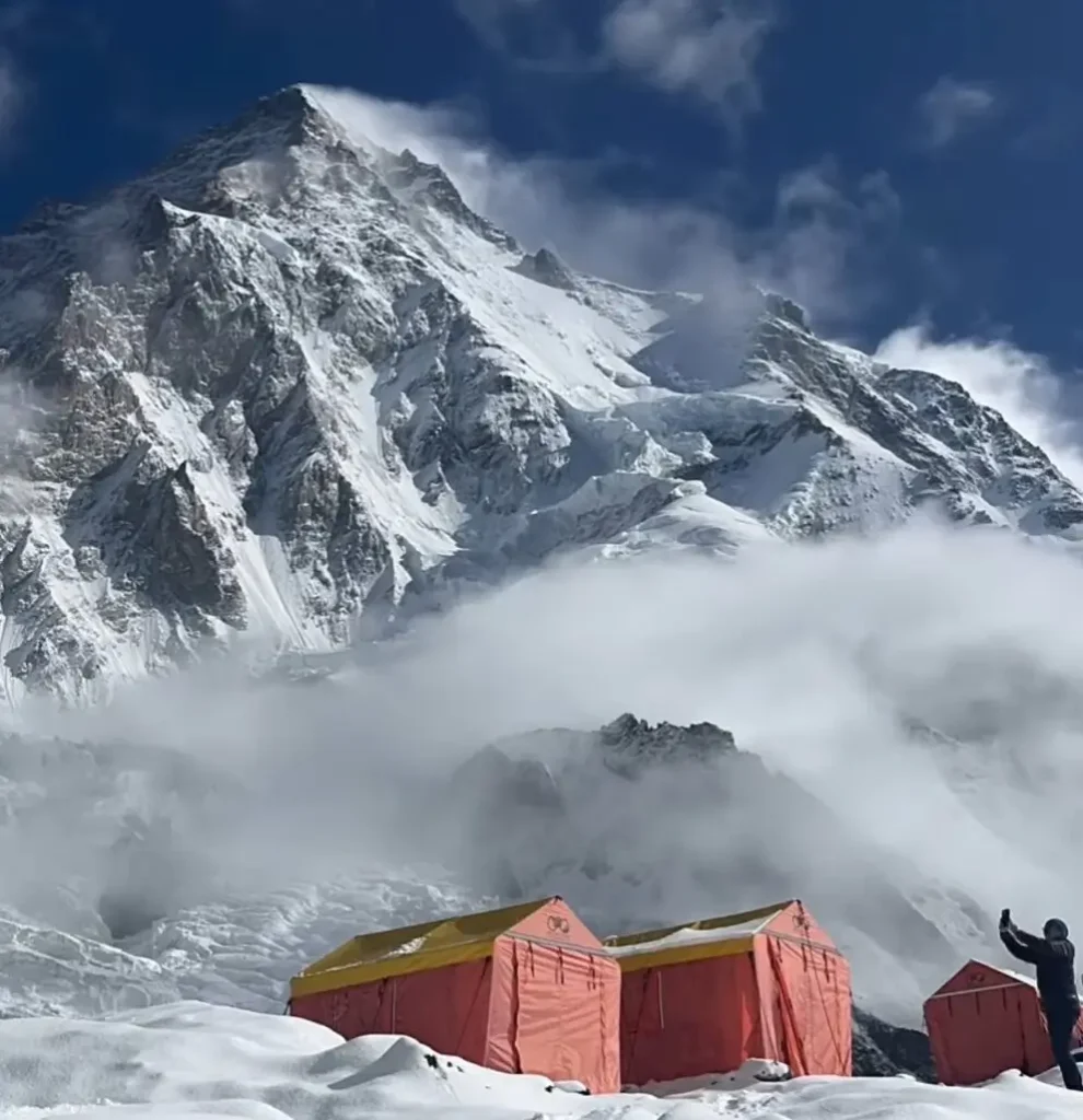 A stunning view of a snow-covered mountain peak with a cloudy sky above. In the foreground, there are red and yellow tents set up on a snowy surface, with a person taking a photo near them.