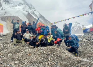 A group of climbers posing for a photo on a rocky terrain with snow-covered mountains in the background. They are wearing colorful outdoor gear and backpacks, with prayer flags strung above them.