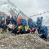 A group of climbers posing for a photo on a rocky terrain with snow-covered mountains in the background. They are wearing colorful outdoor gear and backpacks, with prayer flags strung above them.