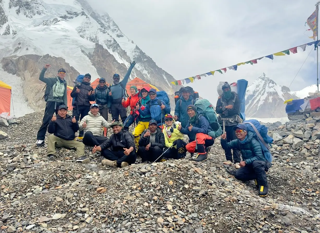A group of climbers posing for a photo on a rocky terrain with snow-covered mountains in the background. They are wearing colorful outdoor gear and backpacks, with prayer flags strung above them.