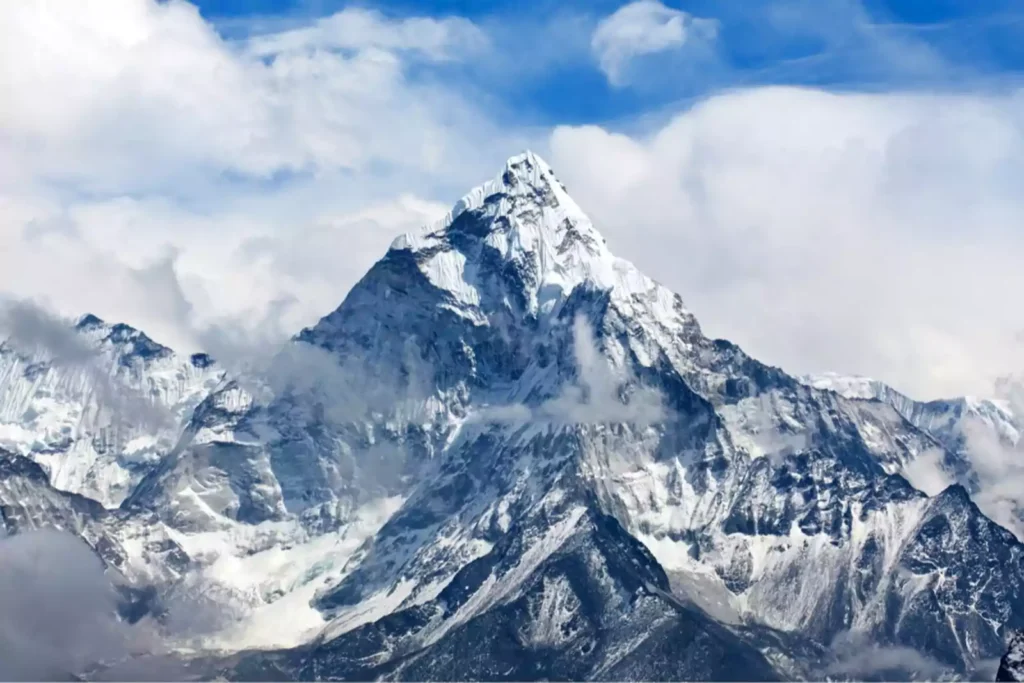 A majestic snow-covered mountain peak rises sharply against a backdrop of a blue sky with scattered clouds. The rugged slopes and ridges of the surrounding mountains are partially shrouded in mist, highlighting the grandeur of the central peak.