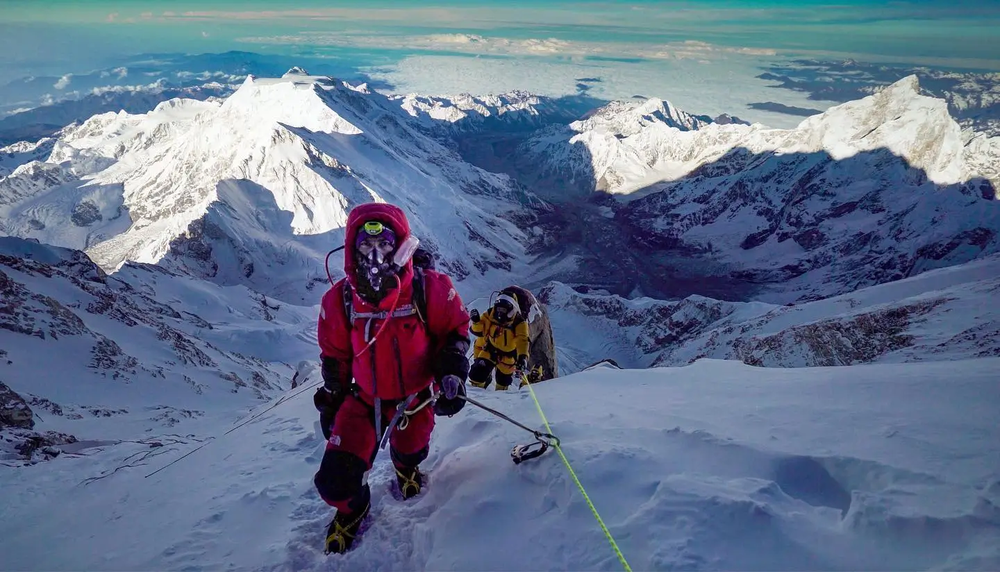 A mountaineer in a pink jacket climbs a snowy ridge, secured with a yellow rope. Another climber in a yellow jacket follows behind. The vast expanse of snow-covered mountains stretches out under a clear sky, with distant peaks and valleys visible.