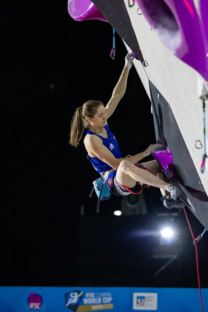 A female rock climber in a blue tank top and gray climbing shoes is ascending an indoor climbing wall with purple holds, secured by a safety rope. She is mid-movement, gripping the holds with chalked hands. The background is dark with bright lights, and a banner at the bottom displays "IFSC World Cup" branding.