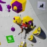 A rock climber in a red shirt and black shorts is scaling an indoor climbing wall with colorful holds in purple, yellow, and silver. The climber is secured with a safety rope and is positioned mid-ascent, gripping and stepping on the holds. The wall features logos for "I Feel Slovenia," "otpbanka," and "Trango," indicating sponsorships or event branding.