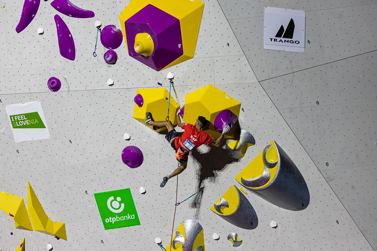 A rock climber in a red shirt and black shorts is scaling an indoor climbing wall with colorful holds in purple, yellow, and silver. The climber is secured with a safety rope and is positioned mid-ascent, gripping and stepping on the holds. The wall features logos for "I Feel Slovenia," "otpbanka," and "Trango," indicating sponsorships or event branding.