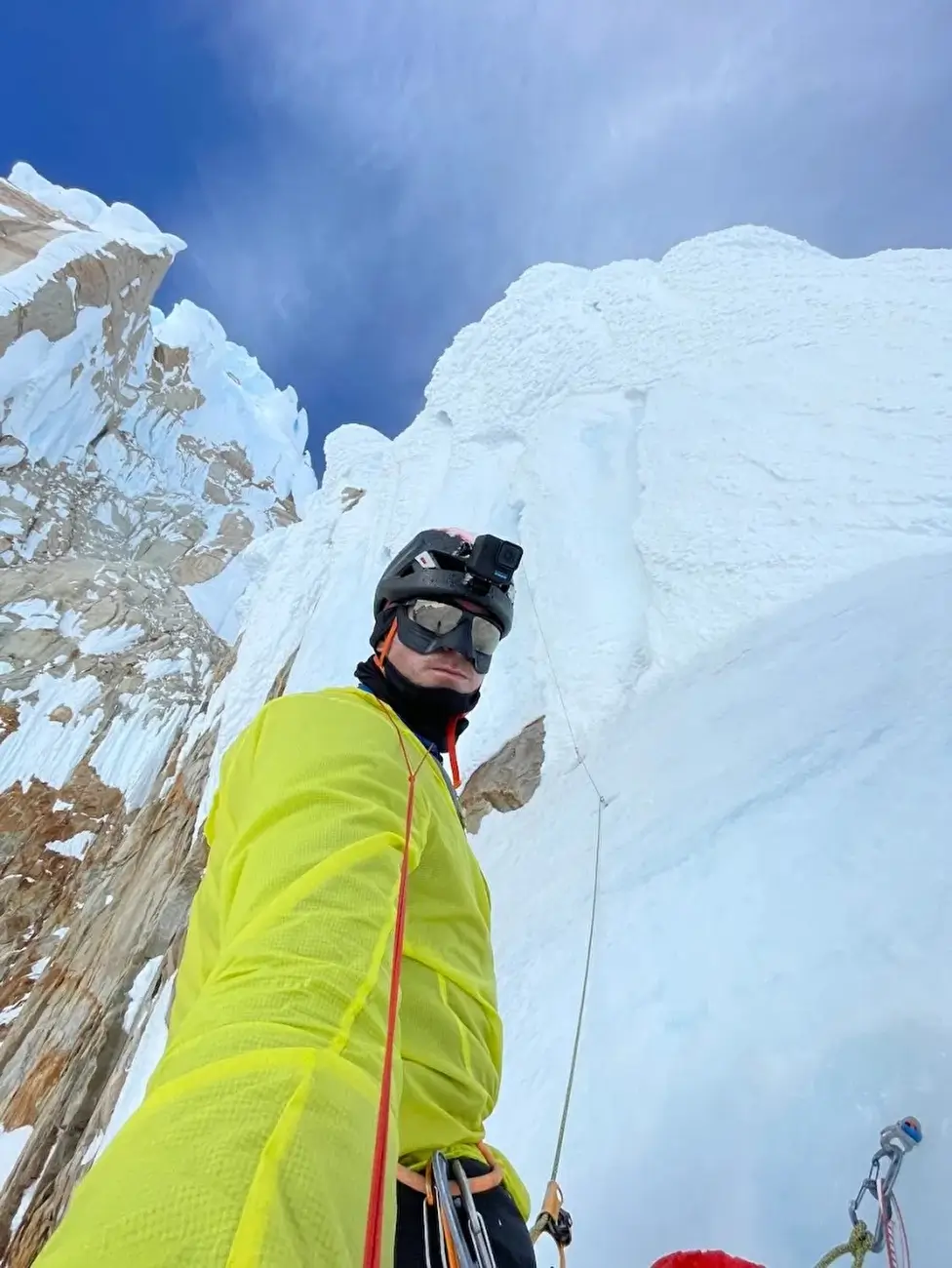 A person in a bright yellow jacket and black helmet with a headlamp is climbing a steep, snow-covered mountain face. He is secured with red and green climbing ropes, with carabiners and gear attached to their harness. The surrounding area features rugged, icy rock formations under a partly cloudy blue sky.
