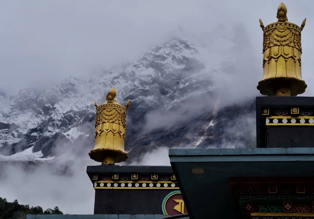 Two ornate golden stupas with intricate designs sit atop a temple structure, set against a misty backdrop of snow-covered mountains. The temple features colorful decorations and traditional patterns, partially obscured by clouds.