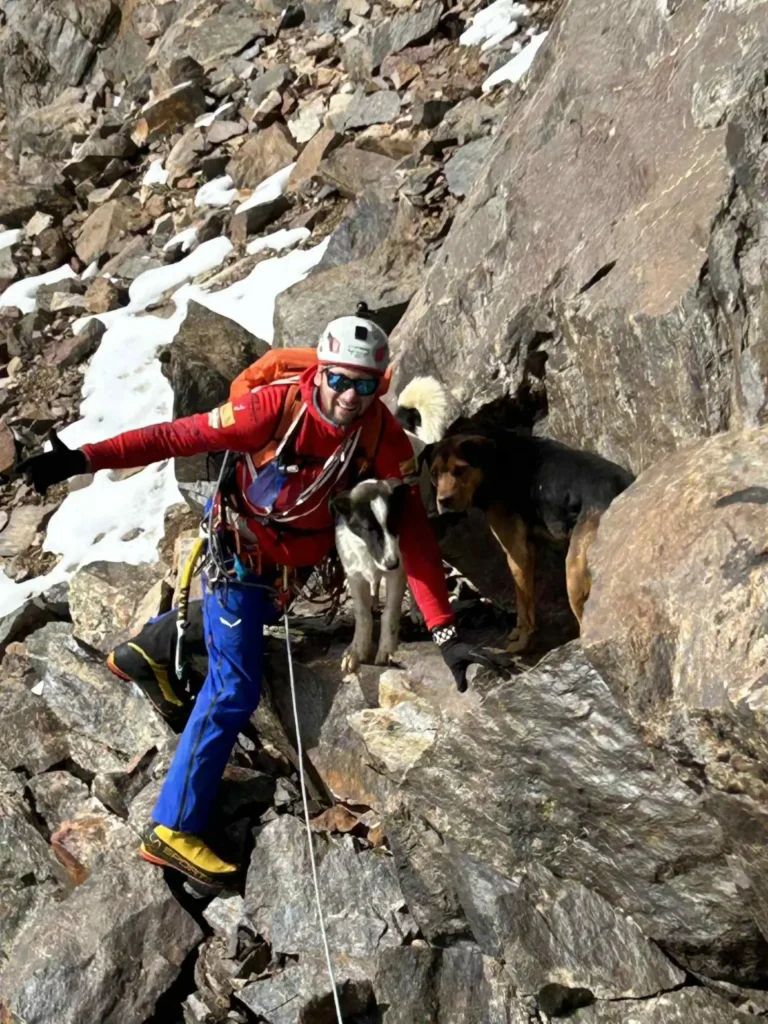 A person in a blue jacket and climbing gear navigates a rocky, snowy mountain slope, accompanied by a white and black dog, with rugged terrain and distant mountains visible under a cloudy sky.