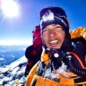 A mountaineer in an orange jacket and headlamp smiles at the camera, with a clear blue sky and bright sun in the background. Snow-covered peaks and another climber in a red jacket are visible, with oxygen equipment attached to the mountaineer's gear.