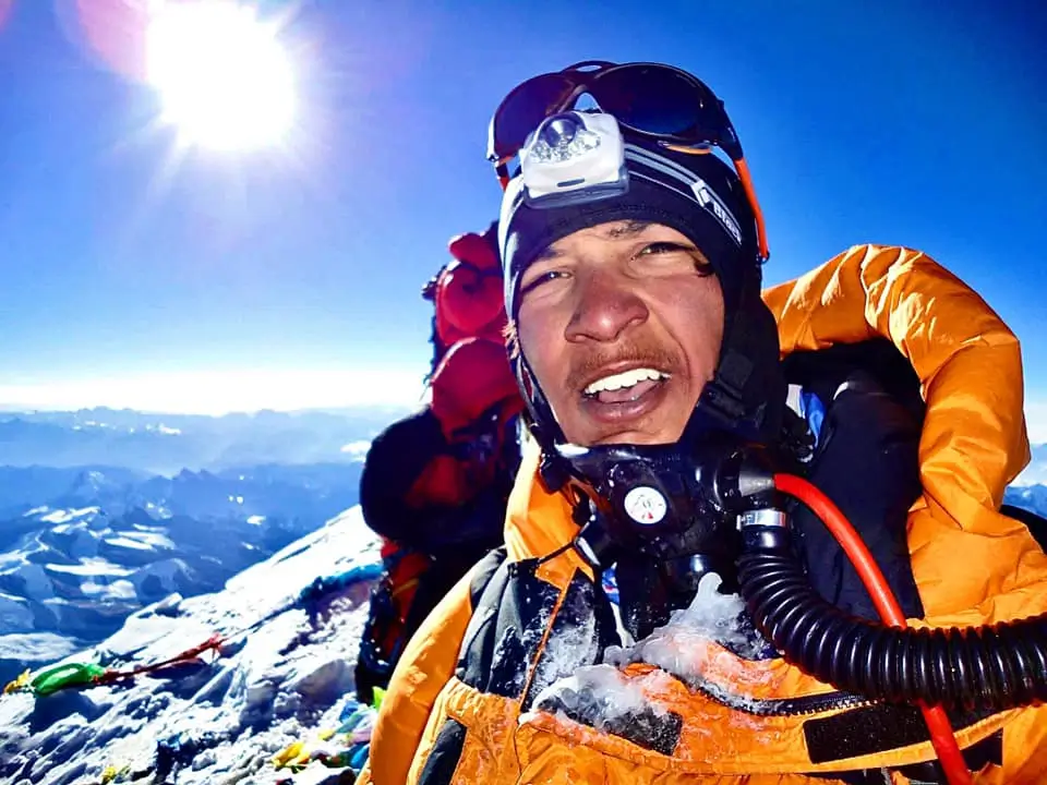A mountaineer in an orange jacket and headlamp smiles at the camera, with a clear blue sky and bright sun in the background. Snow-covered peaks and another climber in a red jacket are visible, with oxygen equipment attached to the mountaineer's gear.