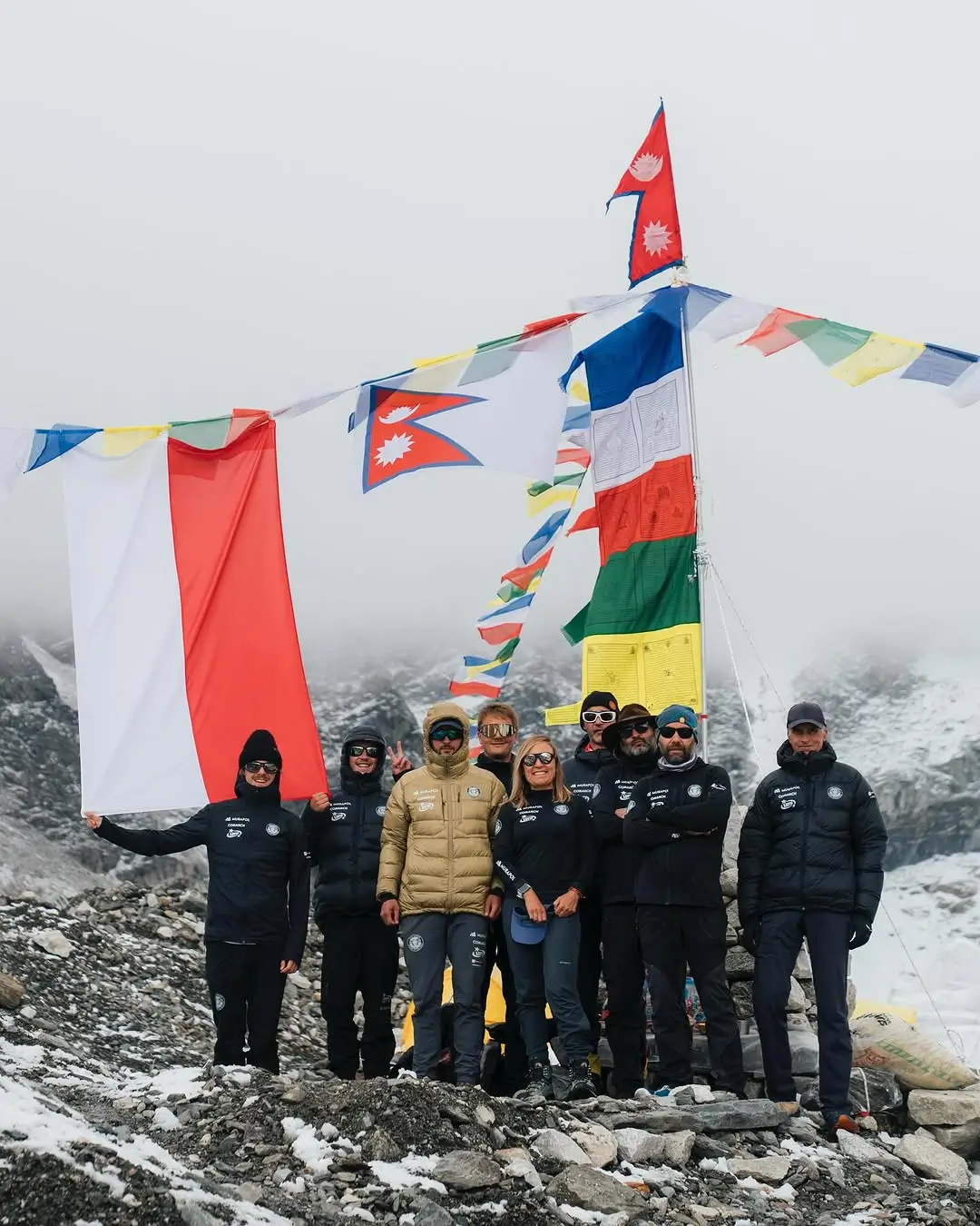 A group of climbers stands on a rocky, snow-covered mountain ridge, holding and surrounded by colorful prayer flags and national flags, including the Nepalese flag. The individuals are dressed in winter mountaineering gear, with some wearing sunglasses and hats. The background features a misty, mountainous landscape, suggesting a high-altitude location.