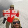Two hikers in winter gear on a snowy mountain ridge, one holding a Georgian flag, with a misty background and a panoramic view of snow-covered peaks under a clear blue sky.