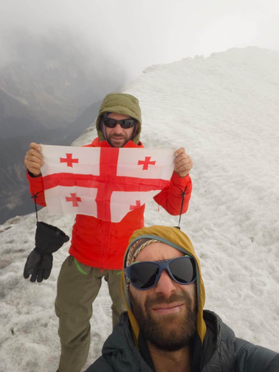 Two hikers in winter gear on a snowy mountain ridge, one holding a Georgian flag, with a misty background and a panoramic view of snow-covered peaks under a clear blue sky.
