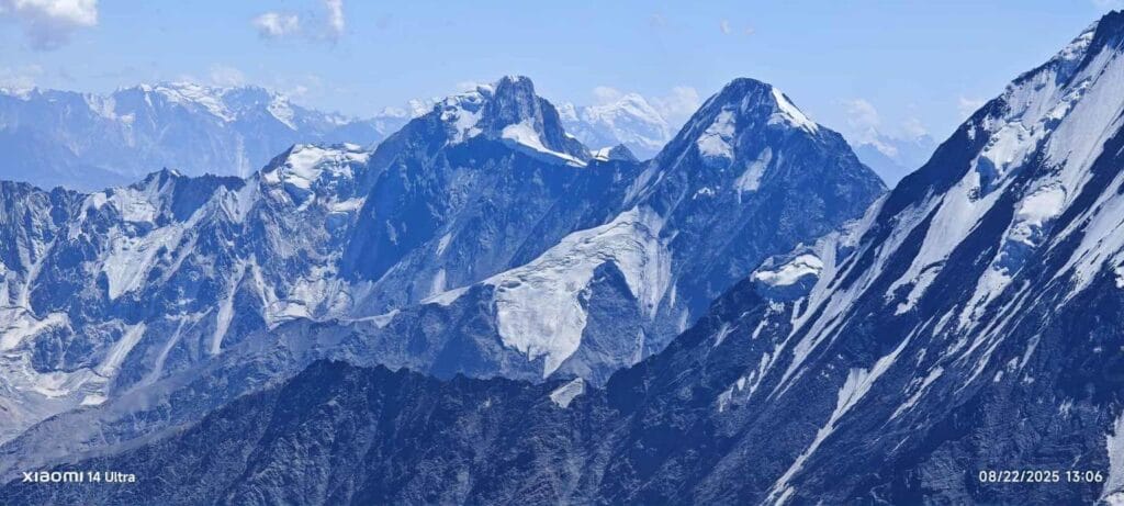 A panoramic view of a rugged mountain range with snow-covered peaks under a clear blue sky, captured on a bright afternoon with some scattered clouds.