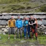 Four hikers stand in front of a stone wall with slate slabs and colorful prayer flags, set against a grassy mountainous backdrop. They are dressed in outdoor gear, including backpacks, trekking poles, and colorful jackets, with one person carrying an additional bag on the ground.