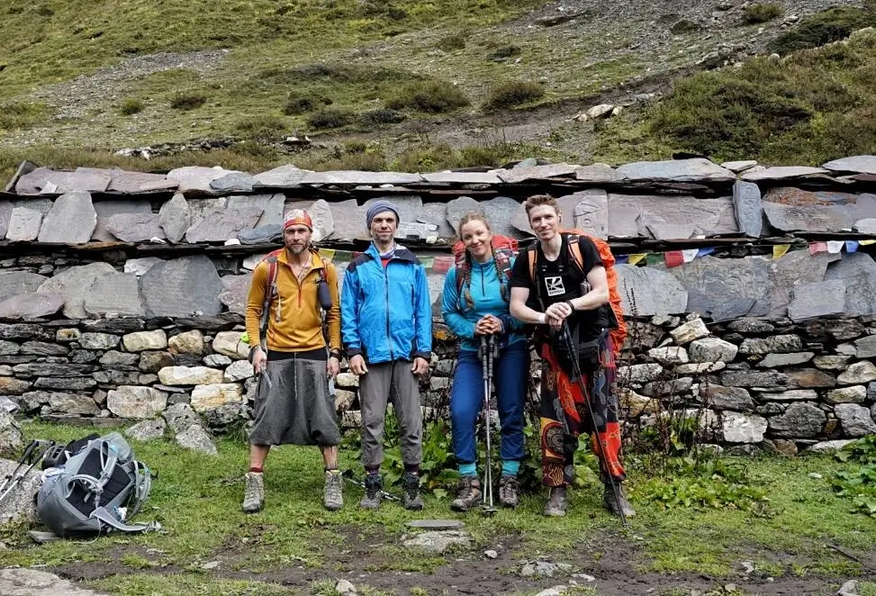 Four hikers stand in front of a stone wall with slate slabs and colorful prayer flags, set against a grassy mountainous backdrop. They are dressed in outdoor gear, including backpacks, trekking poles, and colorful jackets, with one person carrying an additional bag on the ground.
