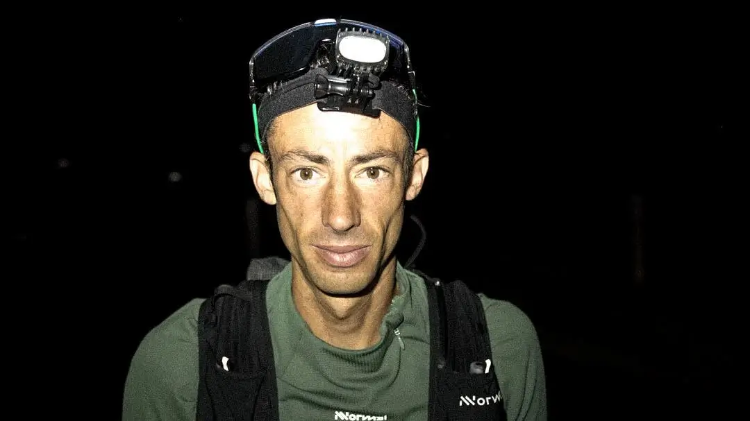 A person wearing a headlamp and a green shirt with the word "Normal" and a logo on it, standing against a dark background.