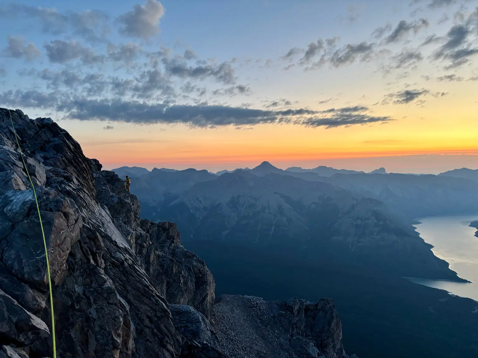 A breathtaking view of a mountain ridge at sunset, with a climber secured by a yellow rope on a rocky cliff. The scene features a vibrant orange and pink sky, scattered clouds, and a range of distant mountains under a serene twilight glow