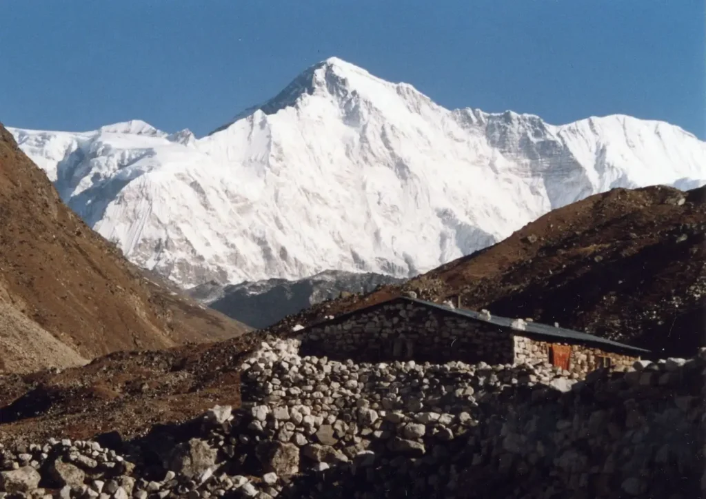 A snow-capped mountain peak towers against a clear blue sky, surrounded by rugged brown hills. In the foreground, a small stone house with a red door and a metal roof sits amidst rocky terrain.