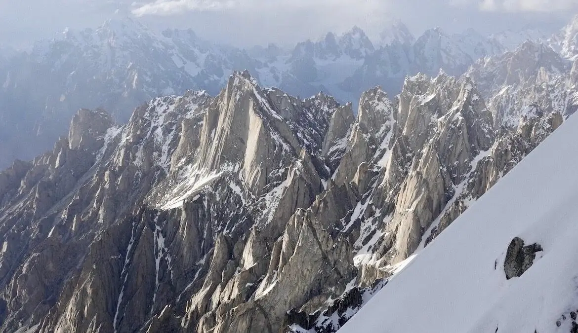 A breathtaking view of a rugged mountain range with sharp, snow-covered peaks under a cloudy sky, captured from a snowy slope in the foreground.