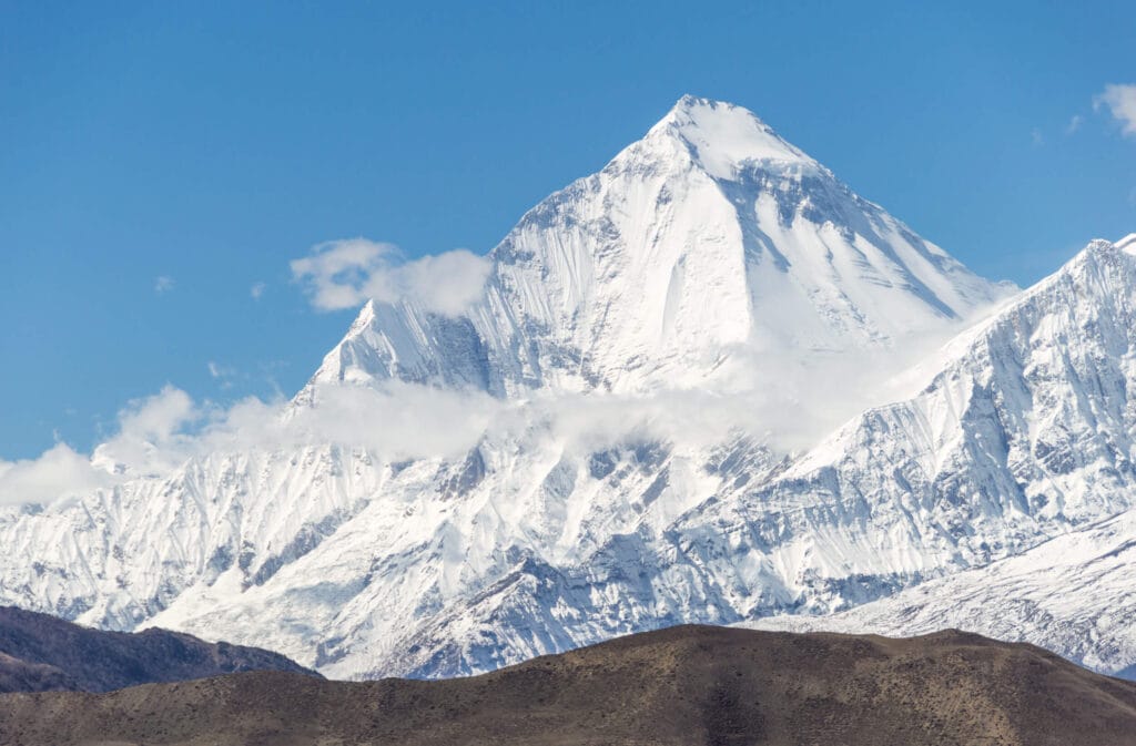 A towering snow-covered mountain peak rises sharply against a clear blue sky, with rugged slopes and ridges partially shrouded by wispy clouds. The foreground features barren, brown hills under the mountain's majestic presence.