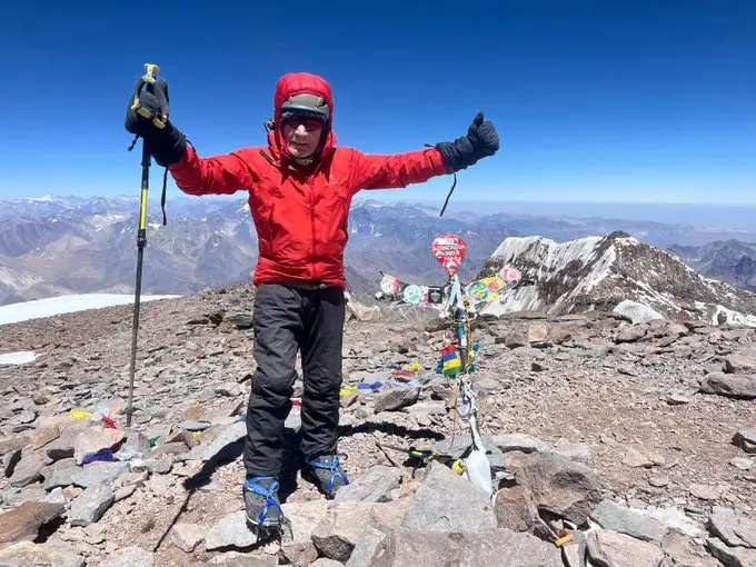 A person in a red jacket and black pants stands on a rocky mountain summit with arms raised, holding trekking poles. A colorful prayer flag pole with decorations stands nearby. Snow-capped mountains and a clear blue sky form a stunning background.
