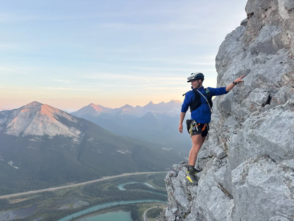 A climber in a blue jacket and helmet ascends a steep rock face, with a stunning backdrop of rugged mountains and a winding river valley below. The early morning or late afternoon light casts a warm hue over the landscape, highlighting the natural beauty of the terrain