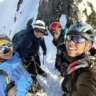 Four climbers wearing helmets and mountaineering gear pose for a selfie on a snowy mountain ridge, with steep snow-covered slopes and rocky cliffs in the background.