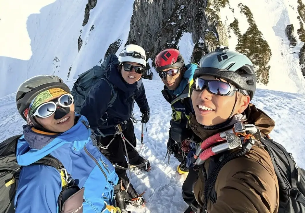 Four climbers wearing helmets and mountaineering gear pose for a selfie on a snowy mountain ridge, with steep snow-covered slopes and rocky cliffs in the background.