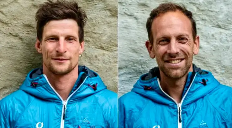 Two individuals wearing blue outdoor jackets with hoods, standing side by side against a stone wall background. The person on the left has short, dark hair and a slight smile, while the person on the right has short, light brown hair and a broader smile.