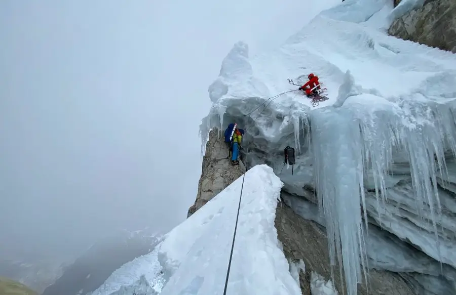 Two climbers are ascending a steep, icy mountain ridge. One climber in a red suit is positioned near the top of an ice wall, secured with ropes and climbing gear. Another climber in a blue suit is lower down, near a rocky outcrop, also roped up. The scene is covered in snow and ice, with icicles hanging from the rock face, and the background fades into a misty, cloudy sky.