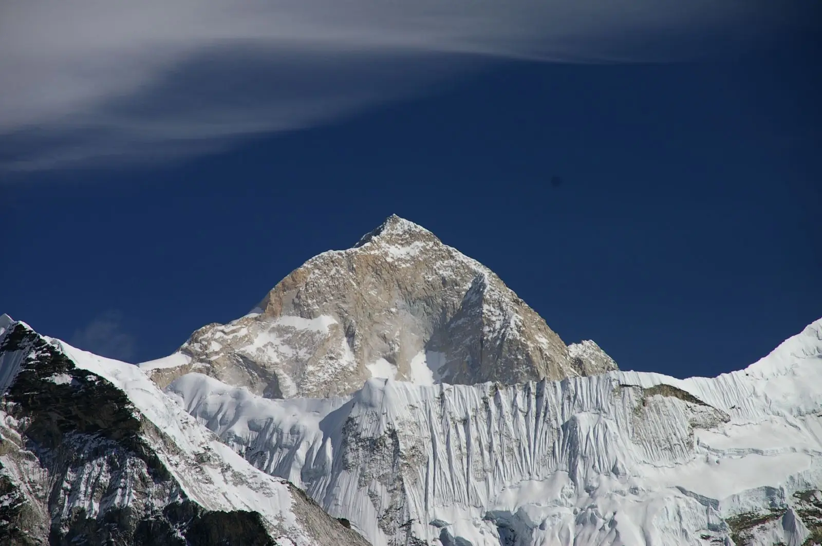 A majestic snow-covered mountain peak rises sharply against a clear blue sky, with rugged slopes and icy ridges prominently displayed, partially shrouded by a thin layer of clouds.