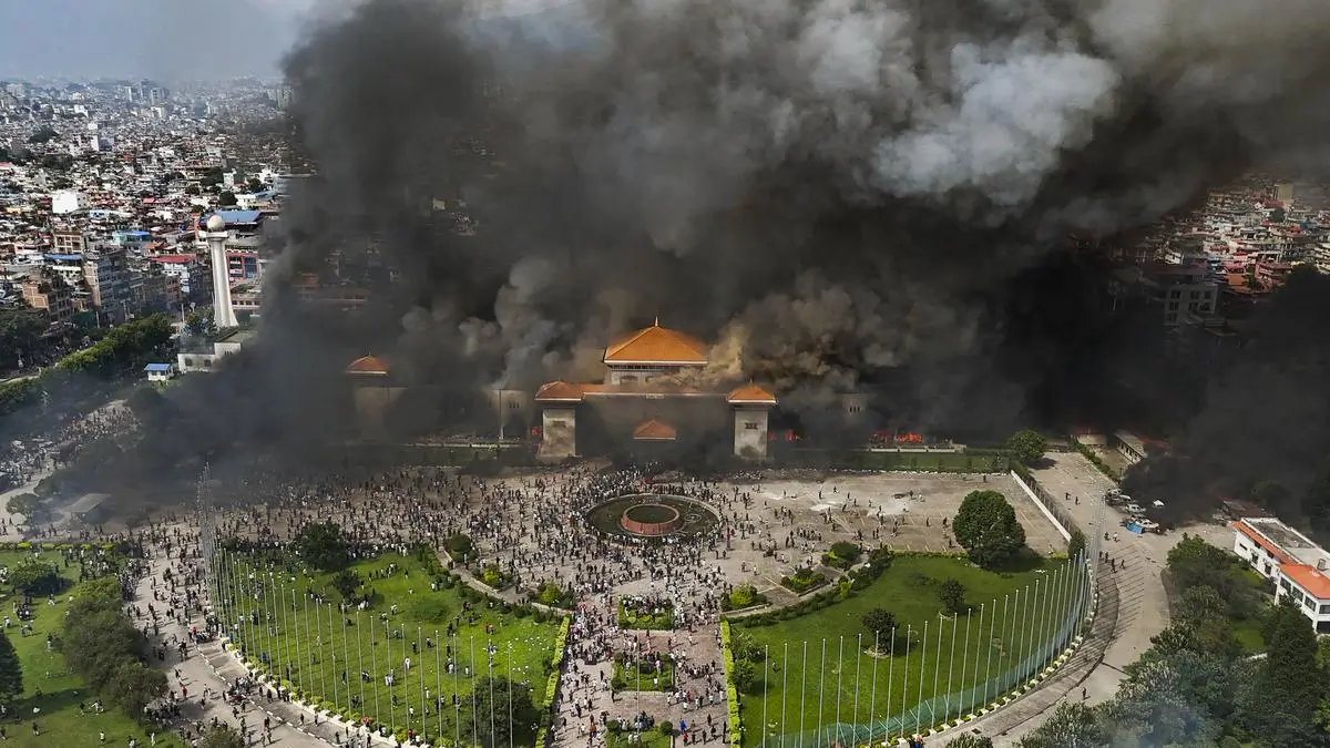 Aerial view of a large public square with a crowd of people gathered, surrounded by green lawns and flagpoles. Thick black smoke rises from a building with an orange roof in the center, indicating a fire or explosion. The surrounding cityscape is visible, with dense buildings extending into the distance.
