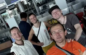 A group of four smiling men posing for a selfie in an airport terminal. They are wearing casual clothing, including t-shirts, a sleeveless top, and backpacks. The background shows check-in counters, screens, and other travelers.