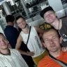 A group of four smiling men posing for a selfie in an airport terminal. They are wearing casual clothing, including t-shirts, a sleeveless top, and backpacks. The background shows check-in counters, screens, and other travelers.