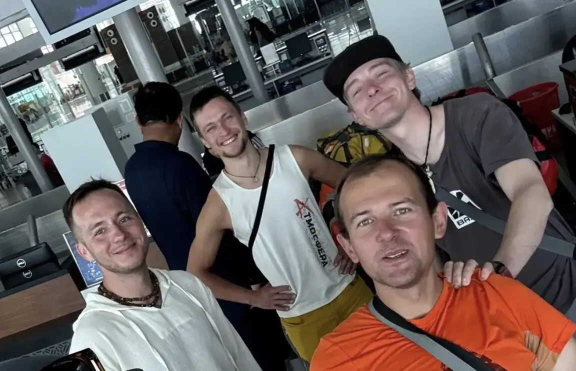 A group of four smiling men posing for a selfie in an airport terminal. They are wearing casual clothing, including t-shirts, a sleeveless top, and backpacks. The background shows check-in counters, screens, and other travelers.