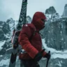 A close-up of a person wearing a red hooded jacket and a headlamp, with skis visible over their shoulder, set against a blurred snowy mountain backdrop.