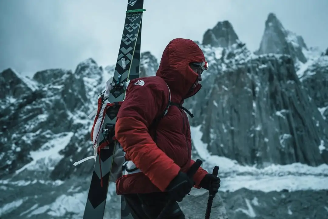 A close-up of a person wearing a red hooded jacket and a headlamp, with skis visible over their shoulder, set against a blurred snowy mountain backdrop.