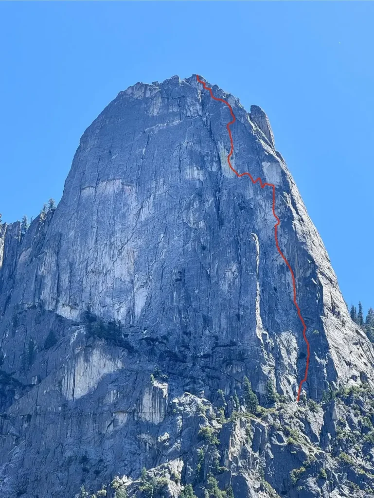 A steep, rocky mountain face under a clear blue sky, with a red line marking a climbing route from the base to the summit.