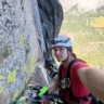 A climber in a red shirt and helmet, equipped with gear, taking a selfie while attached to a rock face with visible climbing equipment and a scenic valley in the background.