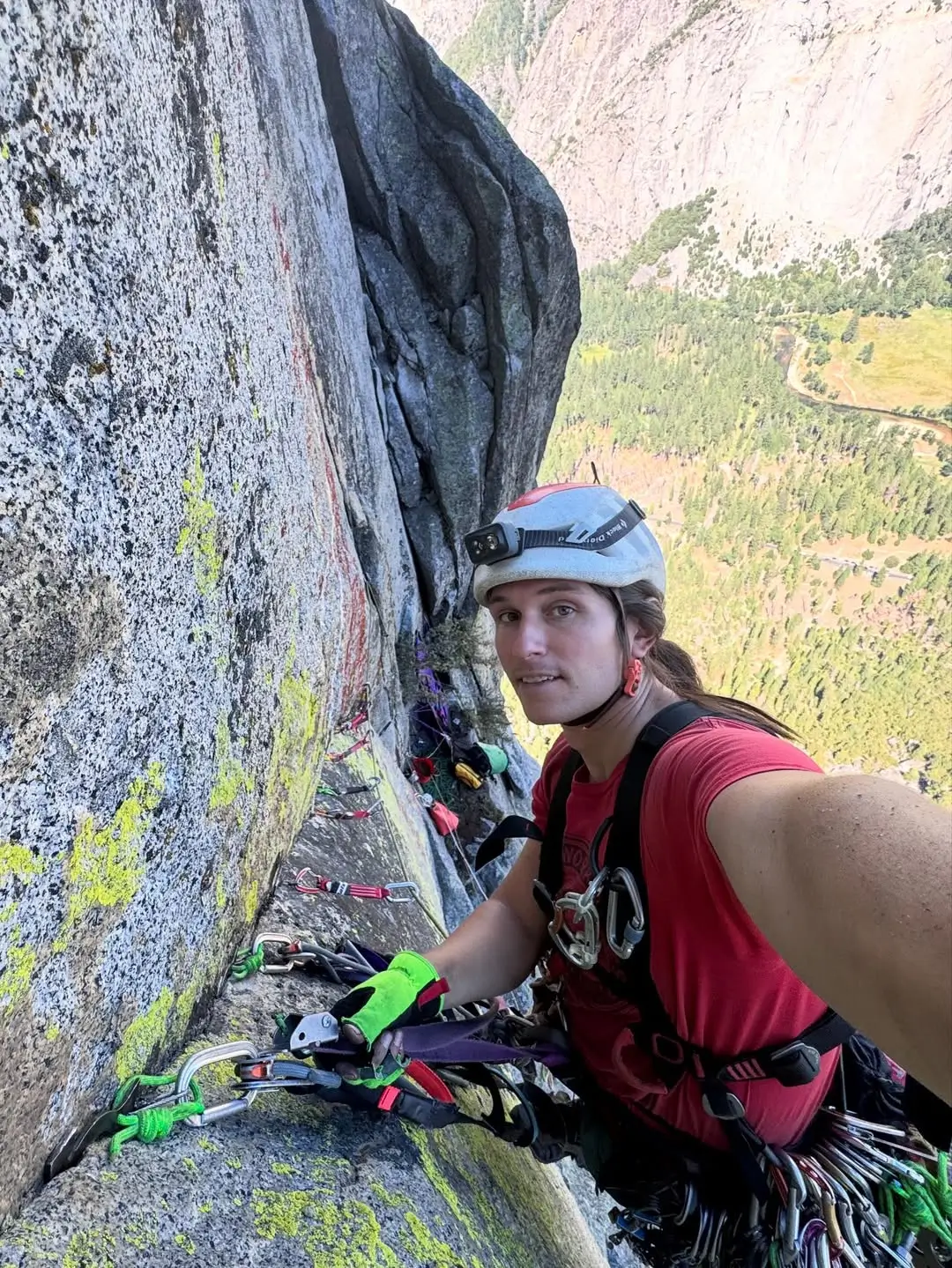 A climber in a red shirt and helmet, equipped with gear, taking a selfie while attached to a rock face with visible climbing equipment and a scenic valley in the background.