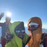 Three climbers in colorful gear take a selfie against a bright blue sky with the sun shining. One person waves, while another holds a camera, with snow-covered mountains faintly visible in the background.