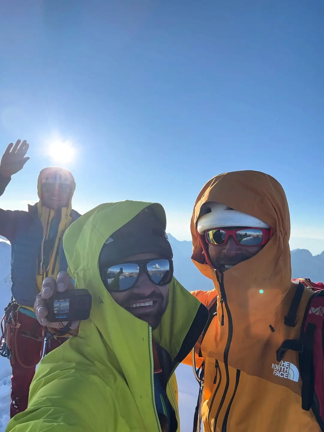 Three climbers in colorful gear take a selfie against a bright blue sky with the sun shining. One person waves, while another holds a camera, with snow-covered mountains faintly visible in the background.