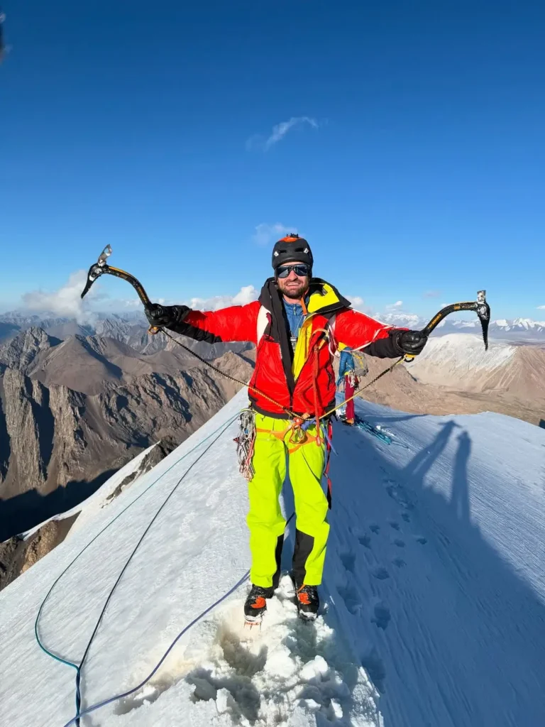 A climber in a red jacket and yellow pants stands triumphantly on a snowy ridge, holding ice axes outstretched, with rugged mountains and a clear blue sky in the background.