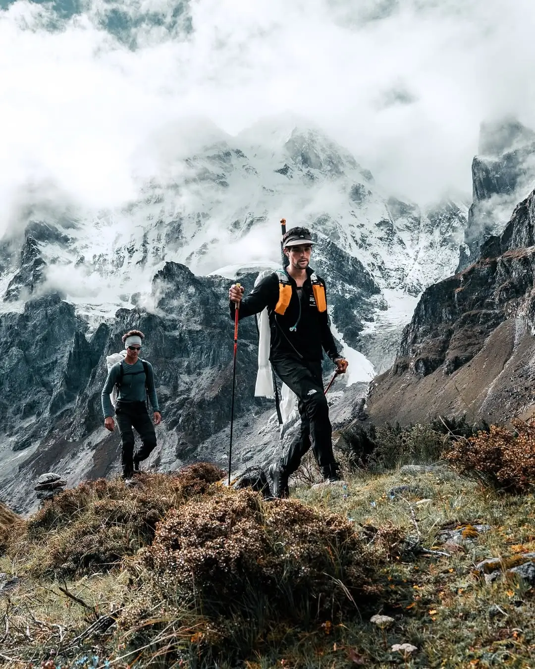 Two hikers with large backpacks stand on a grassy slope, gazing up at a majestic, snow-covered mountain range shrouded in clouds. The text "Le Jannu se montre" is overlaid in the sky.