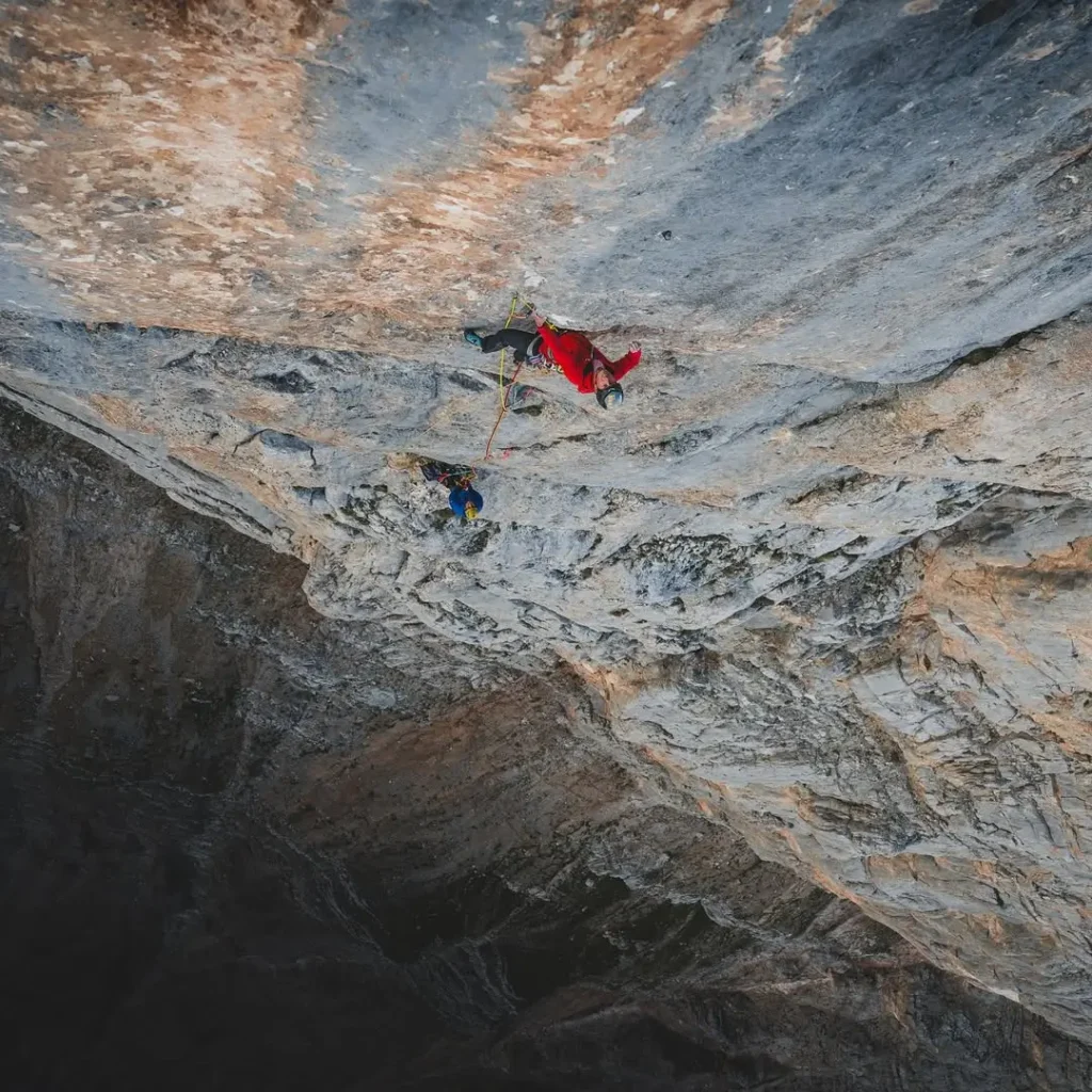 Two climbers on a steep rock face, one in a red outfit and the other in blue, secured with ropes, ascending a challenging section of a mountain cliff under natural light