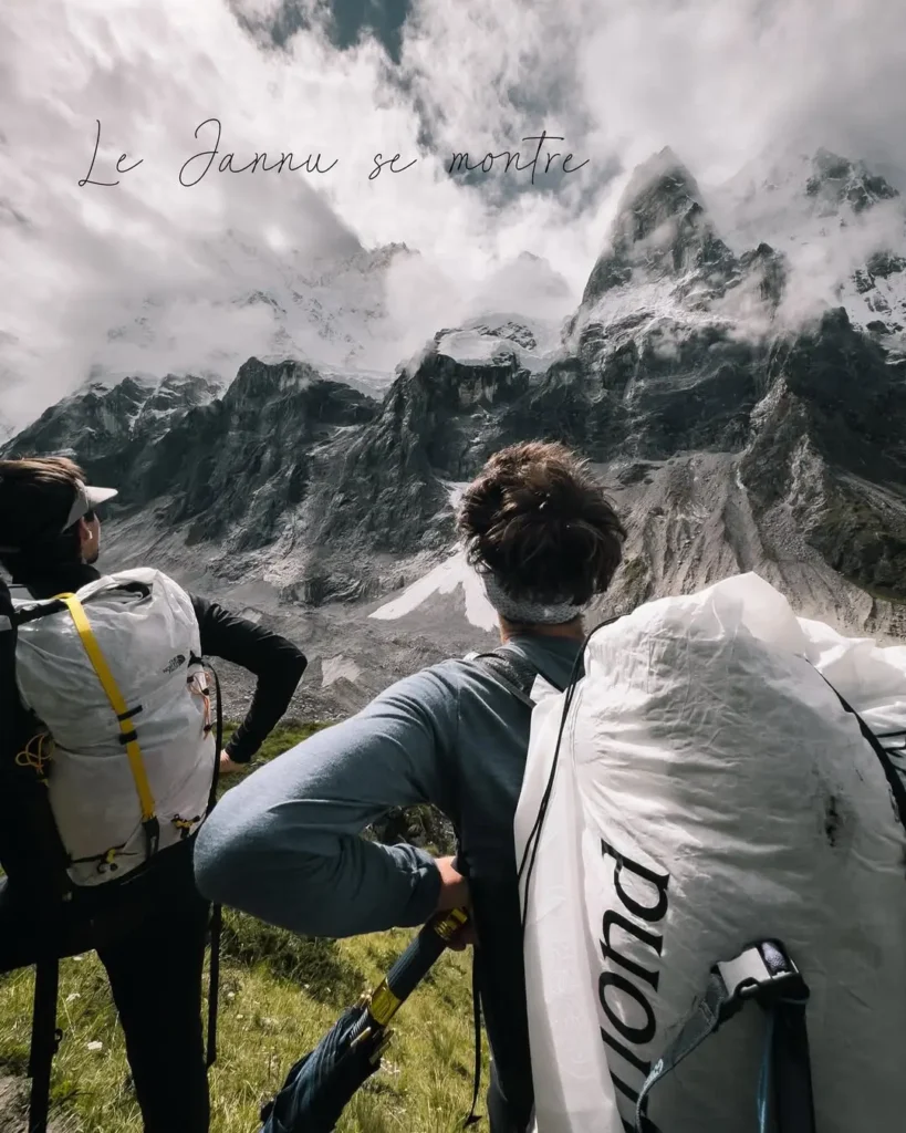 A hiker with a backpack and trekking poles walks across a grassy, rocky terrain with a towering, snow-capped mountain range in the background, partially covered by clouds.