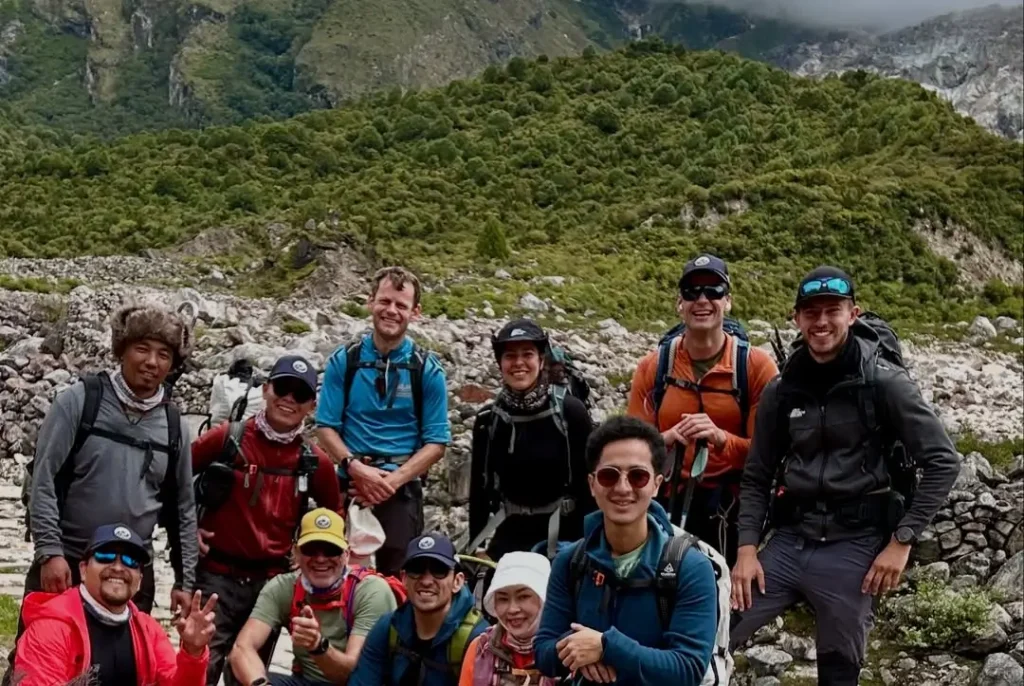 A group of hikers posing for a photo in a mountainous area with lush green hills and rocky terrain in the background. They are wearing backpacks, outdoor clothing, and hats, and some are giving peace signs or smiling. The scene is set against a cloudy sky.