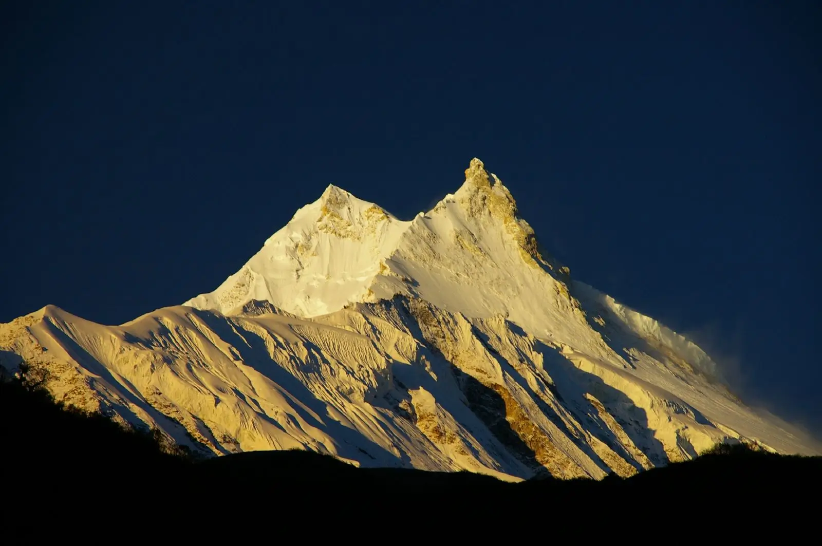 A stunning view of a snow-covered mountain peak illuminated by golden sunlight against a deep blue sky, with rugged slopes and shadows creating a dramatic contrast.